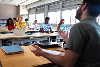 Mann spricht vor einer Klasse Ein Mann spricht zu Frauen und Männern, die vor ihm an Tischen in einem Klassenraum sitzen. Der Mann ist von der Seite fotografiert.